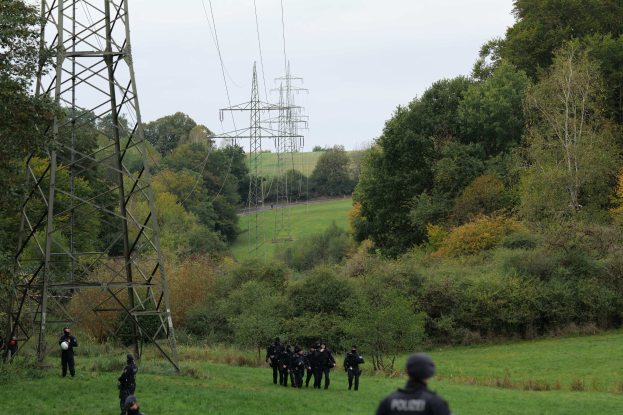 Eine Gruppe von Polizisten geht über ein grünes Feld mit hohen Türmen und Bäumen im Hintergrund unter einem klaren Himmel.