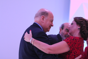 Zwei Menschen umarmen sich warm auf einer Bühne, umgeben von anderen; der Mann trägt ein Armband.