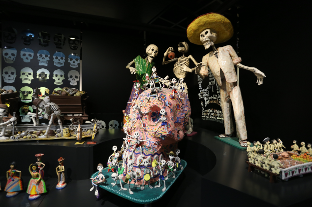 Eine Tag-der-Toten-Ausstellung im Fine Arts Museum in San Francisco, die Spielzeuge, Skelette und andere Gegenstände auf Tischen zeigt, sowie eine Texttafel im Hintergrund in einer dunklen und mysteriösen Atmosphäre.