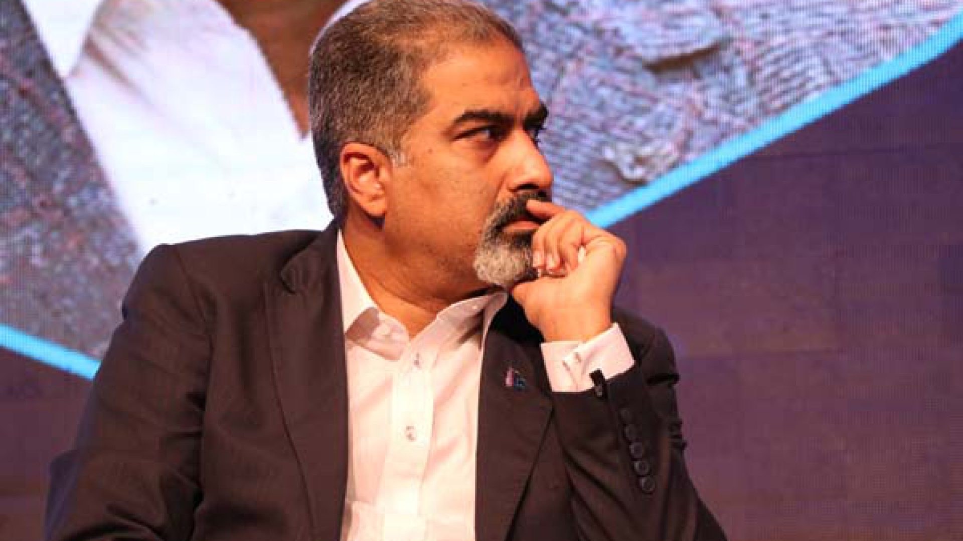 Ein Mann in Anzug und Krawatte sitzt auf einer Bühne vor einem Bildschirm und sieht nachdenklich aus.