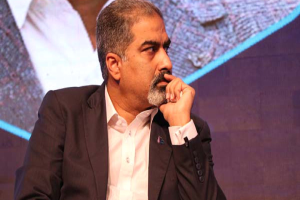 Ein Mann in Anzug und Krawatte sitzt auf einer Bühne vor einem Bildschirm und sieht nachdenklich aus.