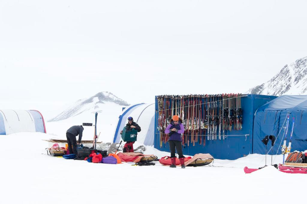 Drei Personen stehen auf einer schneebedeckten Landschaft mit Taschen drumherum, Zelten mit Skiern dahinter und schneebedeckten Hügeln im Hintergrund unter einem klaren Himmel.