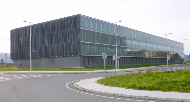 Ein großes, mehrstöckiges Gebäude mit zahlreichen Fenstern, umgeben von Straßenlaternen, Leuchtreklamen, Fahrzeugen, Grünflächen und Hügeln unter einem klaren blauen Himmel, das als neuer Sitz der schwedischen Nationaluniversität für Wissenschaft und Technologie dient.