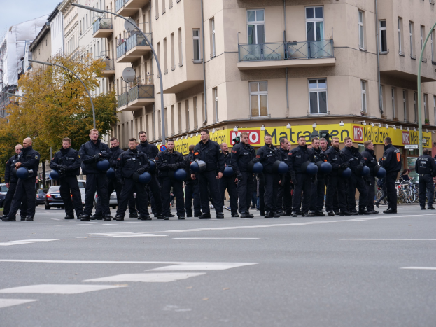 Eine Gruppe von Polizisten in schwarzen Uniformen mit blauen Helmen steht auf einer Straße, die von Laternenpfählen, Bäumen und Glasfenstergebäuden gesäumt ist, unter einem klaren blauen Himmel.