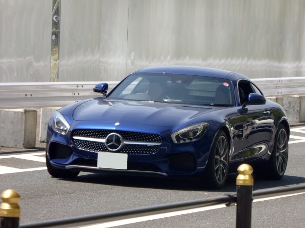 Blauer Mercedes-Benz AMG GT Coupé auf einer Straße mit einer Wand im Hintergrund, mit einem eleganten aerodynamischen Design, eckigen Scheinwerfern, einem großen Kühlergrill, glänzenden großen Rädern und einem luxuriösen Lederinnenraum mit einem modernen Armaturenbrett.