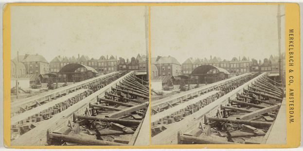 Schwarz-weißes Foto einer Zugschiene mit Gebäuden im Hintergrund, Text 'Merkelbach & Co, Co. Amsterdam' rechts und verstreute hölzerne Gegenstände im Vordergrund.