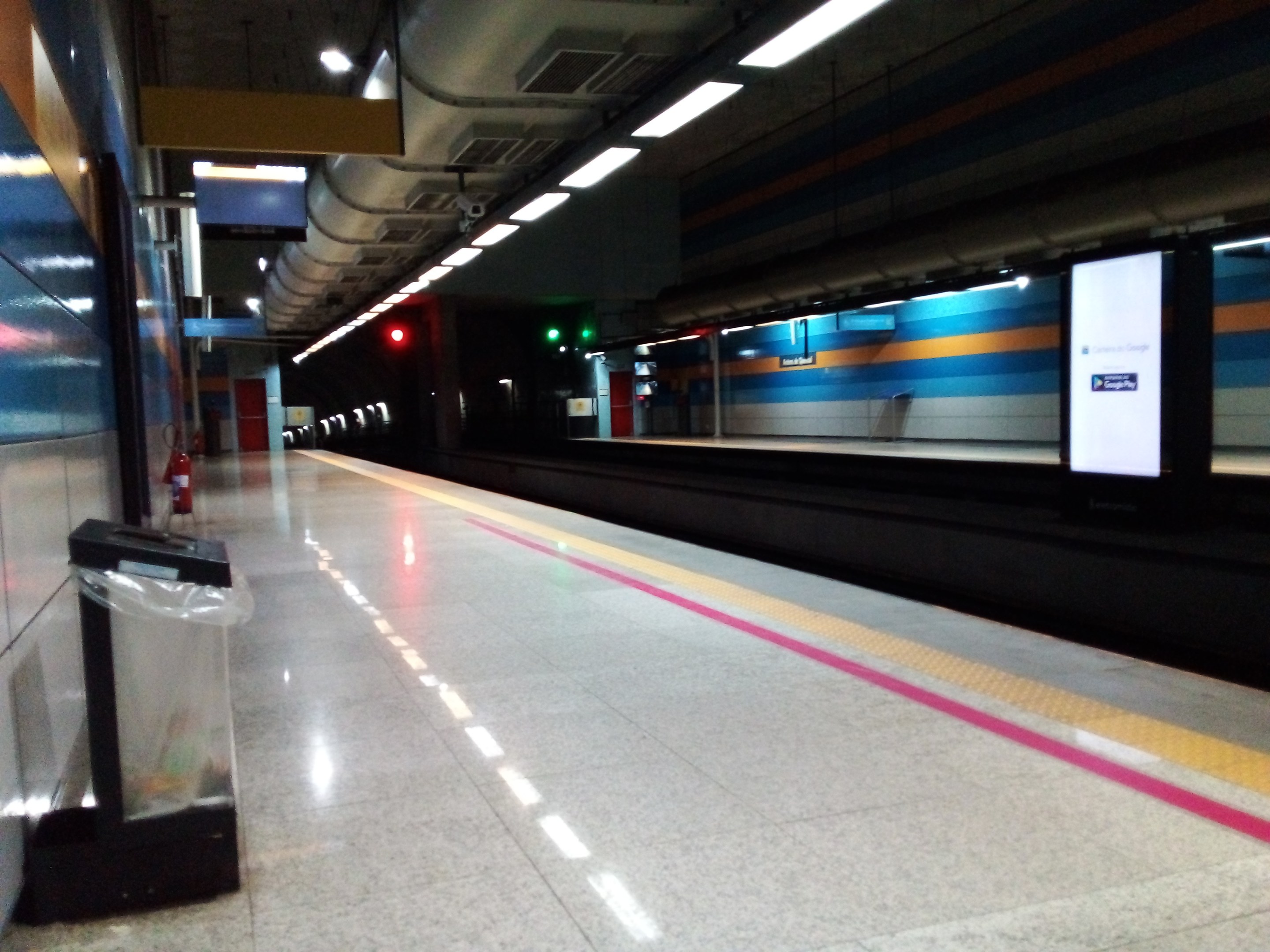 U-Bahn-Station mit einem Zug auf den Schienen, eine Plattform enthält einen Müllkorb, einen Feuerlöscher, Texttafeln, Deckenleuchten und sichtbare Rohre.