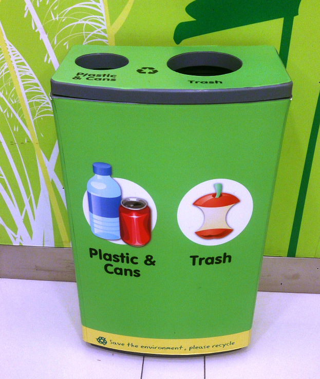 Ein grüner Recycling-Eimer voller Plastik und Dosen steht auf einem weißen Boden, mit einem Plakat im Hintergrund an der Wand.