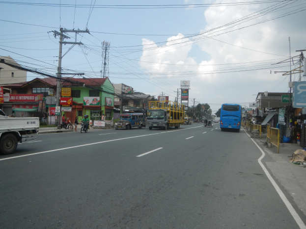 Eine belebte Stadtstraße mit zahlreichen Fahrzeugen, Radfahrern, Gebäuden, Strommasten, Schildern, Bäumen und einem bewölkten Himmel.