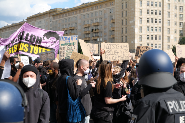 Eine Gruppe maskierter Demonstranten mit Schildern vor einem Gebäude, mit zwei helmträgenden Polizisten rechts daneben, vor einem Hintergrund aus Bäumen und bewölktem Himmel.