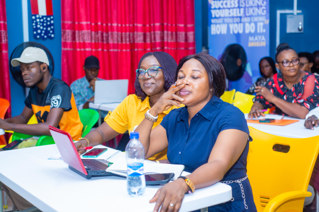 Eine Gruppe von Menschen sitzt um einen Tisch mit Laptops, Flaschen, Mobiltelefonen, Papieren, Stiften und anderen Gegenständen. Im Hintergrund sind eine Wand, ein Vorhang und ein Banner mit Text sowie Fahnen zu sehen.