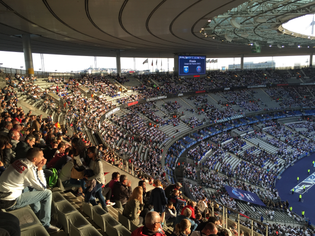 Eine große Menschenmenge sitzt im Allianz Stadion in München, Deutschland, bei einem Fußballspiel, mit einer Bühne auf der rechten Seite, Fahnen, Stangen und einem Bildschirm im Hintergrund und dem Himmel oben sichtbar.