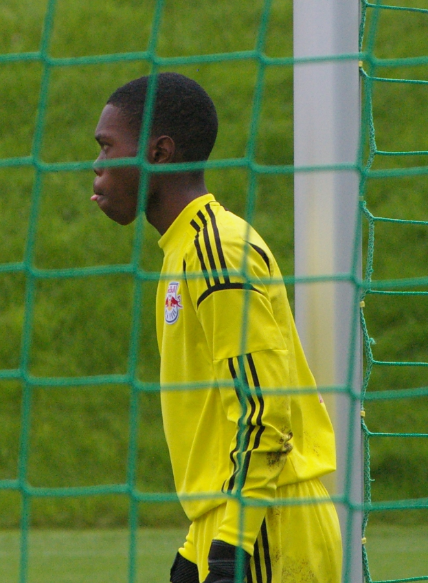 Ein junger Mann in einem gelben T-Shirt und schwarzen Handschuhen steht vor einem Fußballtor, mit saftig grünem Rasen im Hintergrund.