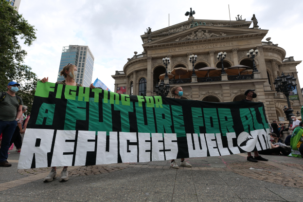 Eine Gruppe maskierter Personen hält ein Transparent mit der Aufschrift "Kämpfen für eine Zukunft für alle Flüchtlinge Willkommen" vor einem Gebäude mit Laternenmasten, Bäumen und einem bewölkten Himmel im Hintergrund.