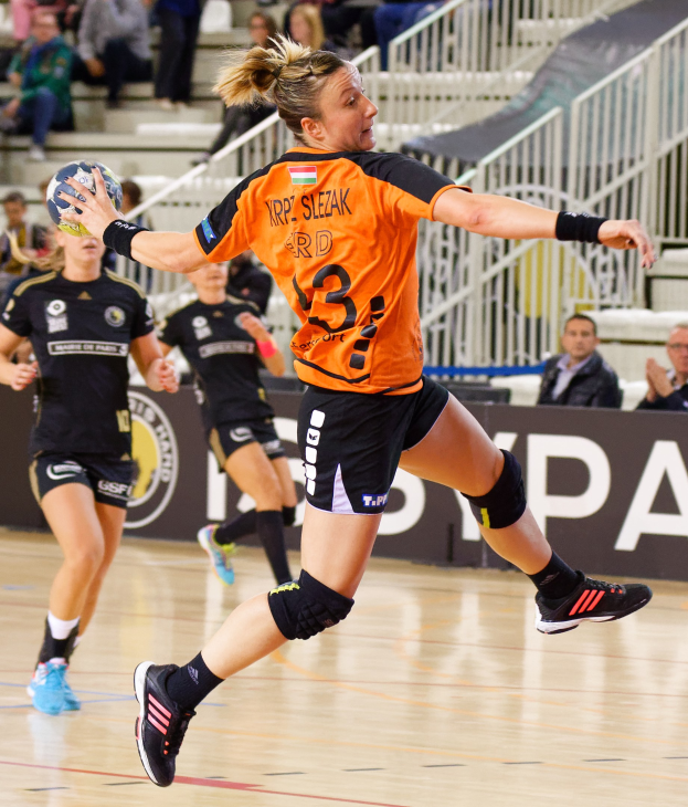 Eine Frau in einem orangefarbenen T-Shirt und schwarzen Shorts hält einen Handball, mit Zuschauern und einem "Krp Slezak - Handball-Weltmeisterschaft 2019"-Schild im Hintergrund.