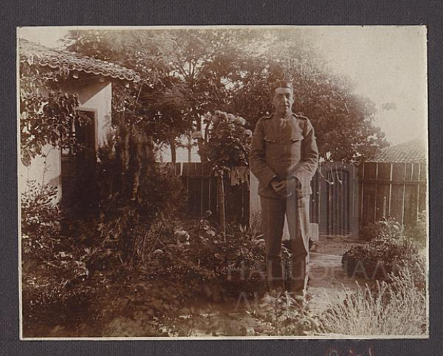 Ein Schwarz-Weiß-Foto eines Mannes in der Uniform eines deutschen Soldaten aus dem Ersten Weltkrieg, der vor einem Haus mit Pflanzen, Bäumen und einem Zaun steht, mit Text am unteren Rand des Fotos.