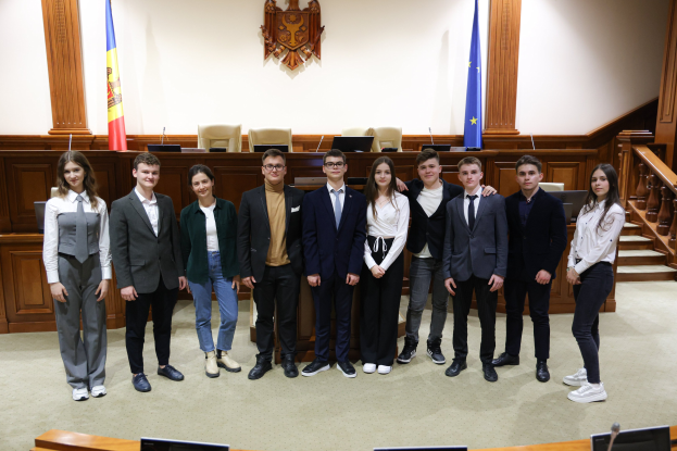 Gruppe von lachenden Menschen, die vor einem Gerichtssaal mit Monitoren und Mikrofonen in der Vordergrund posieren, und eine hölzerne Plattform mit Flaggen und Stühlen im Hintergrund.