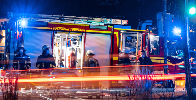 Eine Gruppe von Feuerwehrmännern in Helmen und Schutzausrüstung steht vor einem beleuchteten Feuerwehrauto in der Nacht, mit Pflanzen im Vordergrund, einem Verkehrszeichen auf der rechten Seite und Bäumen im Hintergrund unter einem dunklen Himmel.