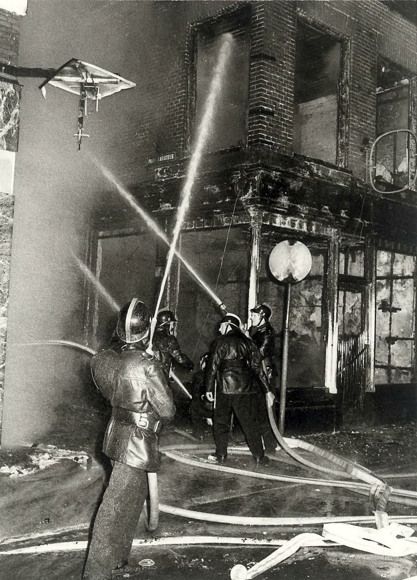 Schwarzes und weißes Bild von Feuerwehrleuten in Helmen mit Schläuchen in den Händen, die vor einem feuerbeschädigten Gebäude stehen.