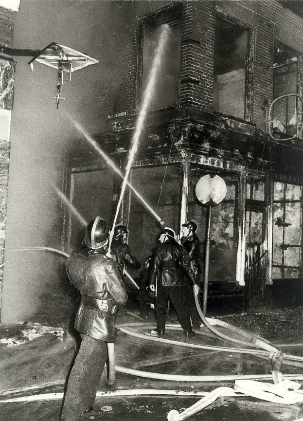 Schwarzes und weißes Bild von Feuerwehrleuten in Helmen mit Schläuchen in den Händen, die vor einem feuerbeschädigten Gebäude stehen.
