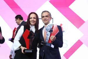 Zwei Menschen auf einer Bühne, die strahlend und Trophäen haltend stehen, mit einem Sichtschutz hinter ihnen.