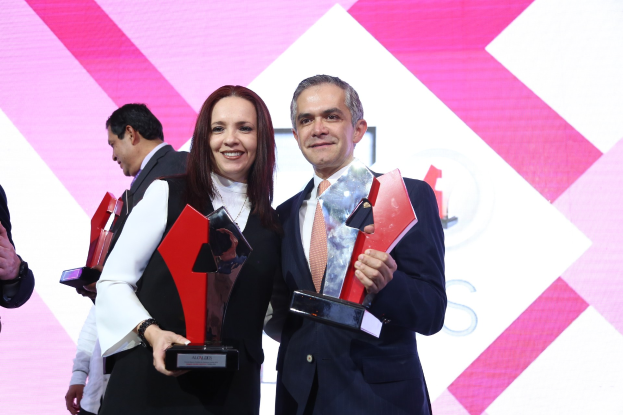 Zwei Menschen auf einer Bühne, die strahlend und Trophäen haltend stehen, mit einem Sichtschutz hinter ihnen.