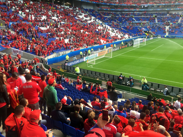 Eine große Menschenmenge in einem Stadion schaut bei einem Fußballspiel zu, mit Menschen auf Sitzen und Stehplätzen, Bannern, einem Torpfosten und Deckenbeleuchtung.