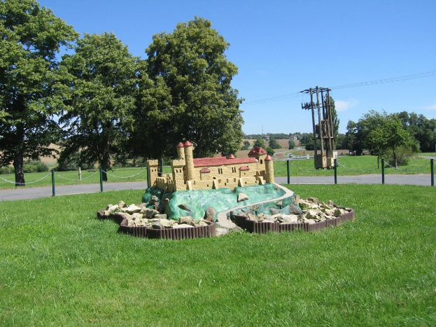 Minigolfplatz mit einem Rasenfeld, umgeben von einem Kettenzaun, Bäumen, Strommasten mit Drähten und einem bewölkten Himmel im Hintergrund.
