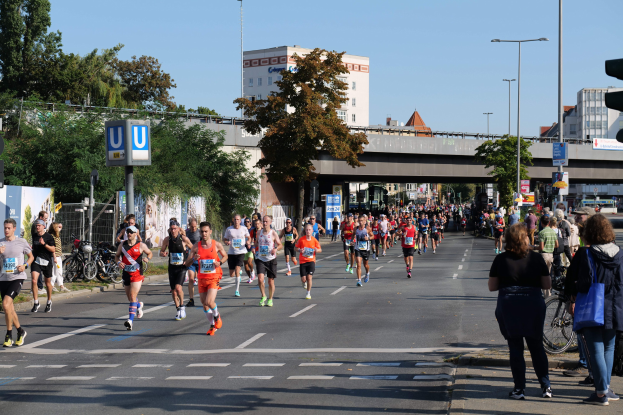 Gruppe von Menschen, die bei einem Marathon auf einer Straße mit Bäumen, Laternen, Schildern, einem Zaun, Fahrrädern und einer Brücke im Hintergrund unter einem klaren blauen Himmel laufen.