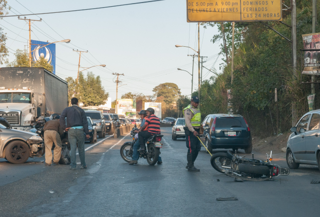 Gruppe von Menschen um ein verunglücktes Motorrad auf der Straße mit mehreren Fahrzeugen, Bäumen, Pfählen, Lampen, Schildern und Himmel im Hintergrund.
