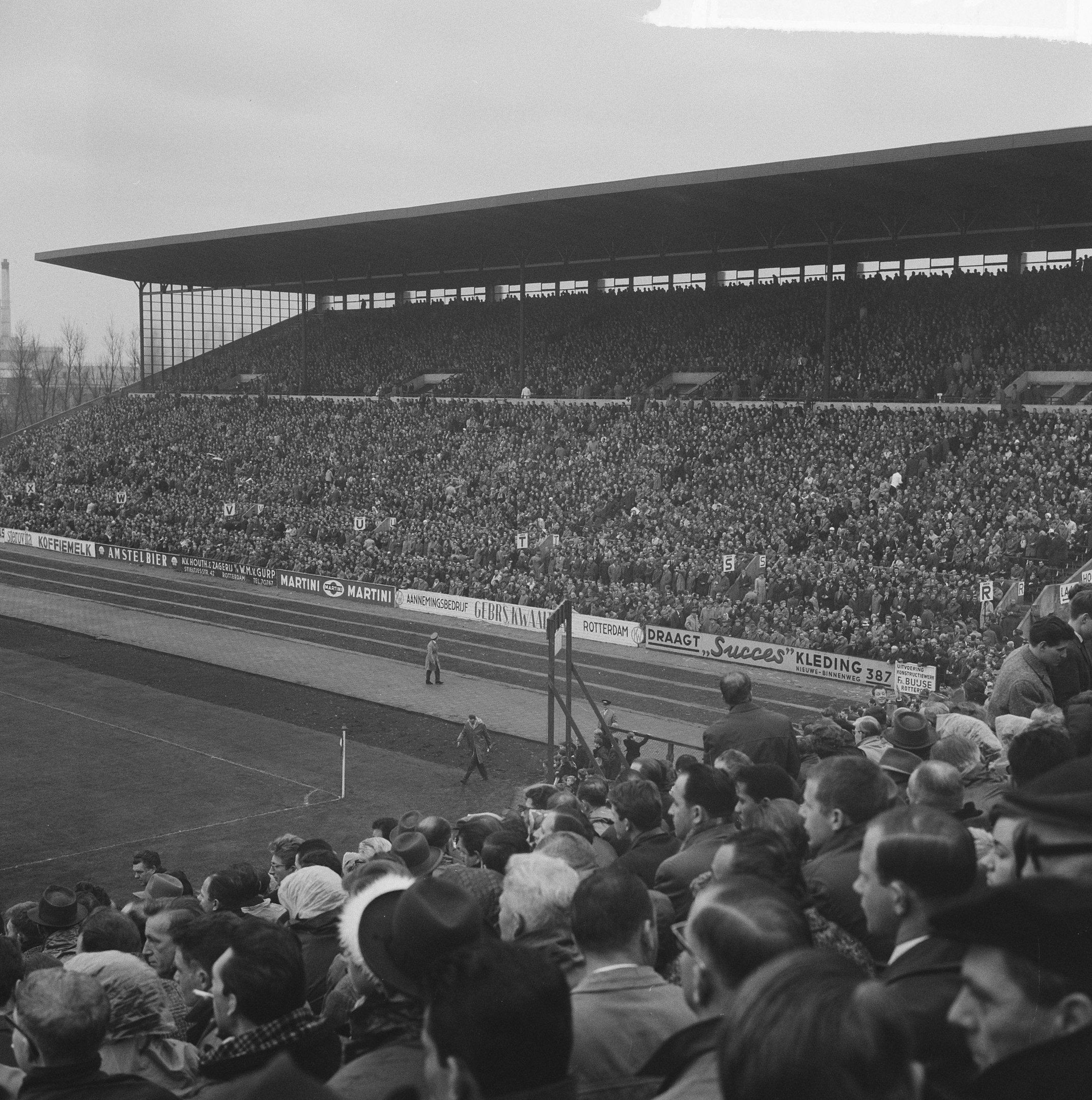 Schwarzes und weißes Foto eines vollen Stadions mit Zuschauern bei einem Fußballspiel, mit Bannern, Pfählen, einem Schuppen, Bäumen, einem Turm und einer bewölkten Himmel