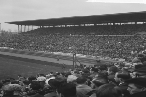 Schwarzes und weißes Foto eines vollen Stadions mit Zuschauern bei einem Fußballspiel, mit Bannern, Pfählen, einem Schuppen, Bäumen, einem Turm und einer bewölkten Himmel