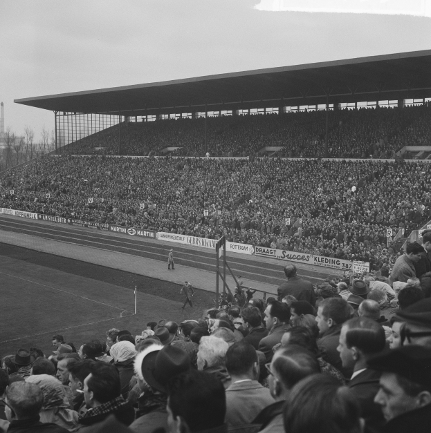 Schwarzes und weißes Foto eines vollen Stadions mit Zuschauern bei einem Fußballspiel, mit Bannern, Pfählen, einem Schuppen, Bäumen, einem Turm und einer bewölkten Himmel