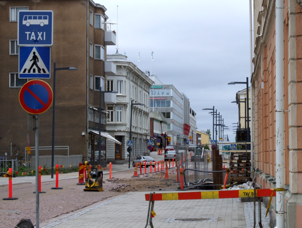 Stadtstraße mit Gebäuden, Straßenmöblierung, Fahrzeugen, einer Baustelle und Bäumen unter einem bewölkten Himmel.
