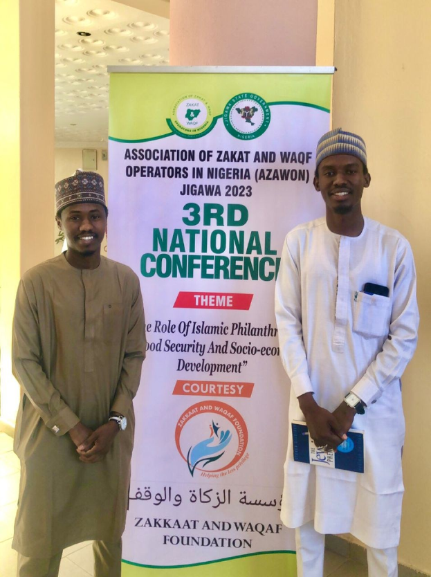 Zwei Männer neben einem Banner '3. Nationaler Kongress' mit einer Säule und Deckenbeleuchtung im Hintergrund.