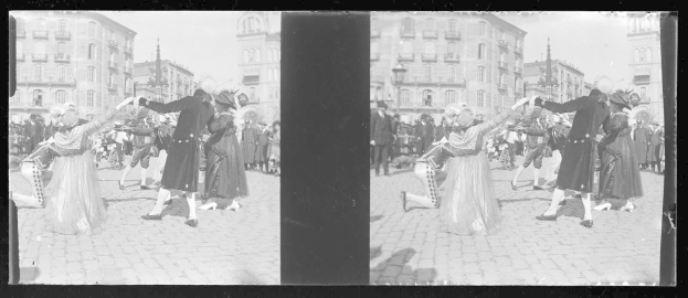 Ein Schwarz-Weiß-Foto einer Gruppe von Menschen, die auf der Straße tanzen, während eines Karnevals-Umzugs, gekleidet in Kostüme, mit Gebäuden, Straßenlaternen und Straßenlaternen im Hintergrund unter einem klaren Himmel.