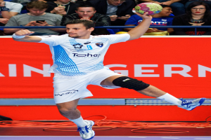 Ein Mann in der Luft mit einem Handball, im Hintergrund sitzen Menschen und ein Banner mit Text.