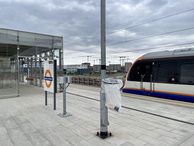 Ein Zug fährt auf einem Bahnsteig ein, vorne ein Pfahl und ein Mülleimer, links ein Schuppen und im Hintergrund Gebäude, Bäume, Pfähle, Drähte und Himmel.