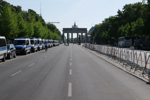 Lange Reihe von Polizeiwagen auf der Straße vor dem Brandenburger Tor in Berlin, Deutschland, mit Menschen auf Fahrrädern und auf der Straße, Barrieren, Bäumen, einem Bogen mit Statuen im Hintergrund und sichtbarem Himmel.