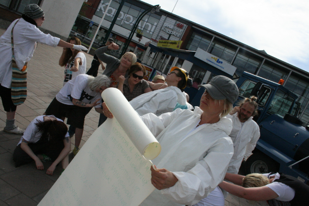 Eine Gruppe von Menschen, einige in Kostümen und Mützen, versammelt sich in der Mitte, während einige zu kämpfen scheinen, während eine Person ein Papier in der Vordergrund hält. Im Hintergrund gibt es Gebäude, Pflanzen, Pfähle und Tafeln mit Text, ein Fahrzeug auf der rechten Seite, einen Weg am Boden und den Himmel oben.