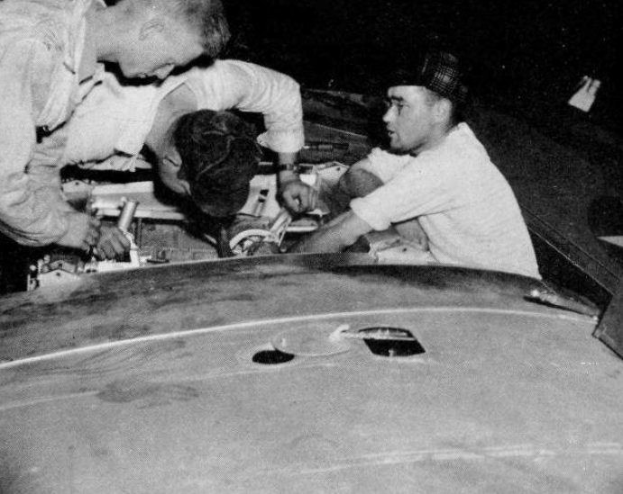 Drei Männer mit Mützen reparieren ein schwarz-weißes Auto in einer Garage umgeben von Werkzeugen und Materialien.