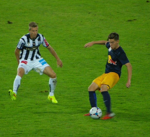 Zwei Männer spielen Fußball auf einem grünen Feld, einer trägt ein blaues Hemd und gelbe Shorts, der andere ein weiß-schwarzes gestreiftes Hemd und weiße Shorts.
