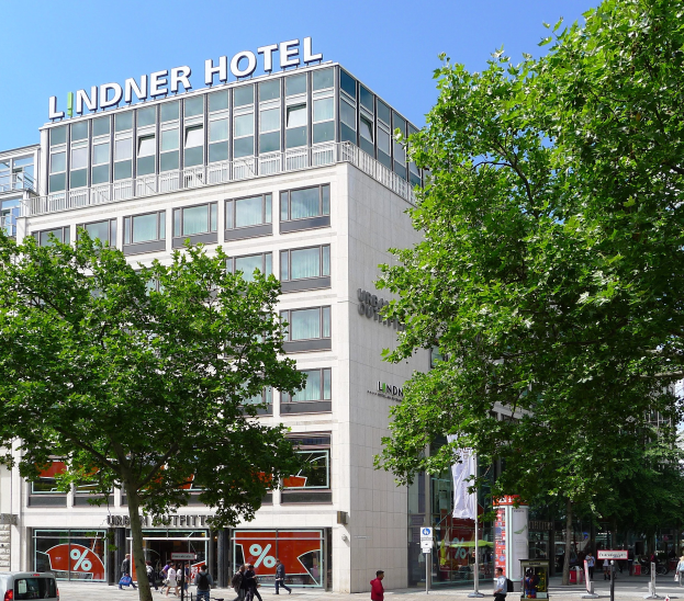 Großes Gebäude mit Glasfront und der Aufschrift "Lindner Hotel" in Berlin, umgeben von Bäumen, Fahrzeugen und Passanten.