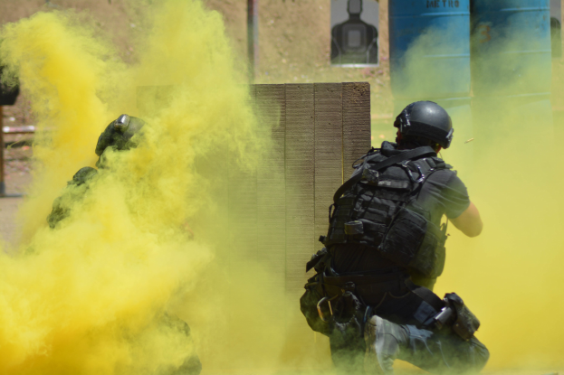 Zwei Polizisten in Einsatzausrüstung mit gelbem Rauch aus dem Mund, der vor einer Wand mit einer Tafel und anderen Gegenständen steht.