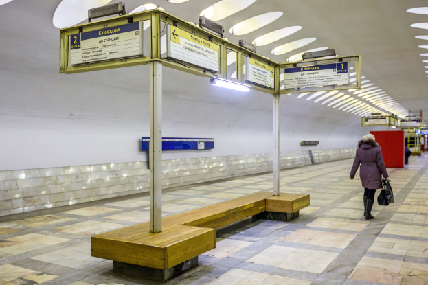 Eine Frau in einem Mantel geht durch eine Moskauer U-Bahn-Station, vorbei an Bänken, Informationsanzeigen und Deckenleuchten, während sie eine Tasche trägt.