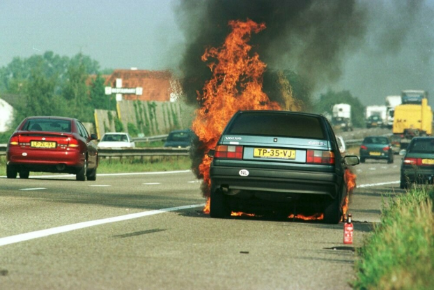 Ein Auto, das in Flammen steht, auf der Straße mit anderen Fahrzeugen in der Nähe, Bäume und Gebäude im Hintergrund bei einem klaren blauen Himmel und Gras mit einem Feuerlöscher auf der rechten Seite.