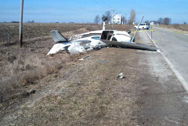 Ein Kleinflugzeug stürzte auf der Seite einer Straße, umgeben von Gras und Pfählen, mit Bäumen, Häusern und einem klaren blauen Himmel im Hintergrund.