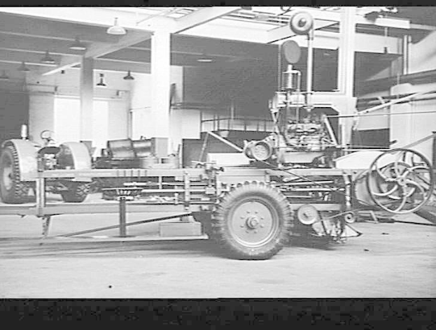 Schwarzes und weißes Foto eines Traktor-Schlepper-Traktors in einer Fabrik mit Säulen, Deckenbeleuchtung und umgeben von Maschinen.