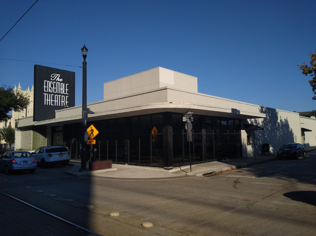 Das Ensemble Theatre in Austin, Texas, mit Fahrzeugen auf der Straße, einer Straßenlaterne und Brettern an Pfählen, Bäumen im Hintergrund und einem blauen und weißen Himmel.