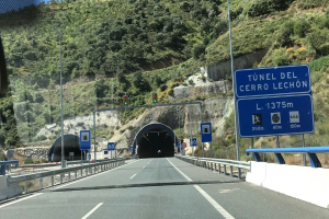 Auto fährt durch einen Tunnel auf einer Straße mit Geländern, Schildern, Pfosten, Verkehrszeichen, Bäumen, Felsen und einem klaren blauen Himmel.